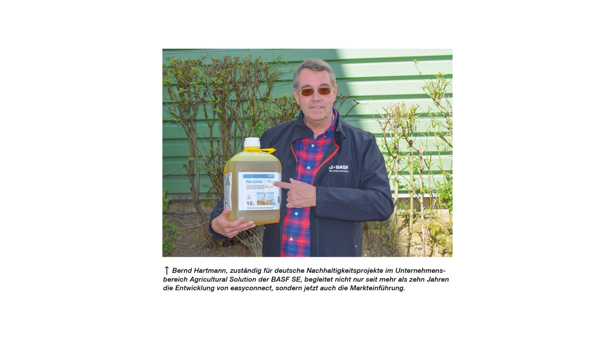 Bernd Hartmann von BASF zeigt einen Kanister mit dem Produkt Revytrex im Außenbereich vor einer grünen Wand und Hecke. 