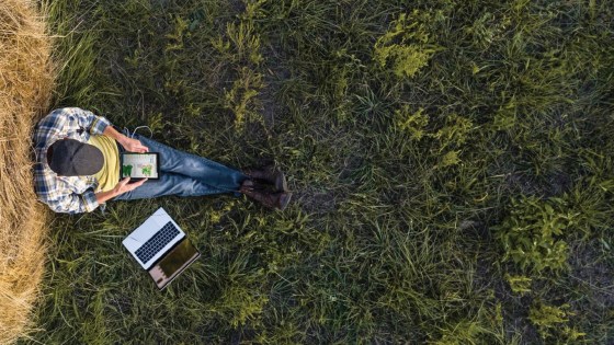 Landwirt sitzt entspannt auf einer Wiese neben einem Laptop und nutzt ein Tablet zur digitalen Feldanalyse.